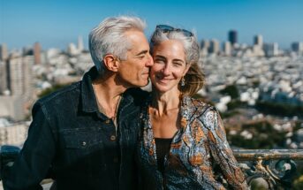 A smiling couple with gray hair stands close together outdoors, with a city skyline and tall buildings blurred in the background. The man kisses the woman’s temple as she smiles at the camera, both enjoying a sunny day.