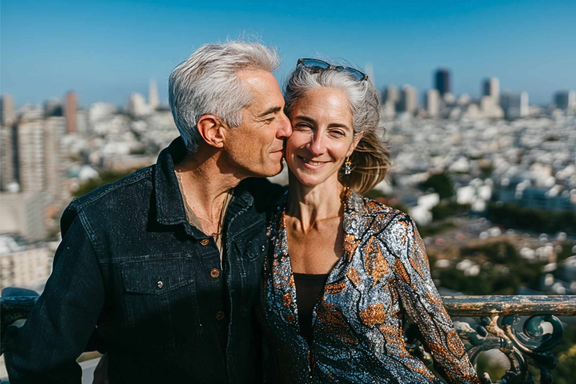 A smiling couple with gray hair stands close together outdoors, with a city skyline and tall buildings blurred in the background. The man kisses the woman’s temple as she smiles at the camera, both enjoying a sunny day.