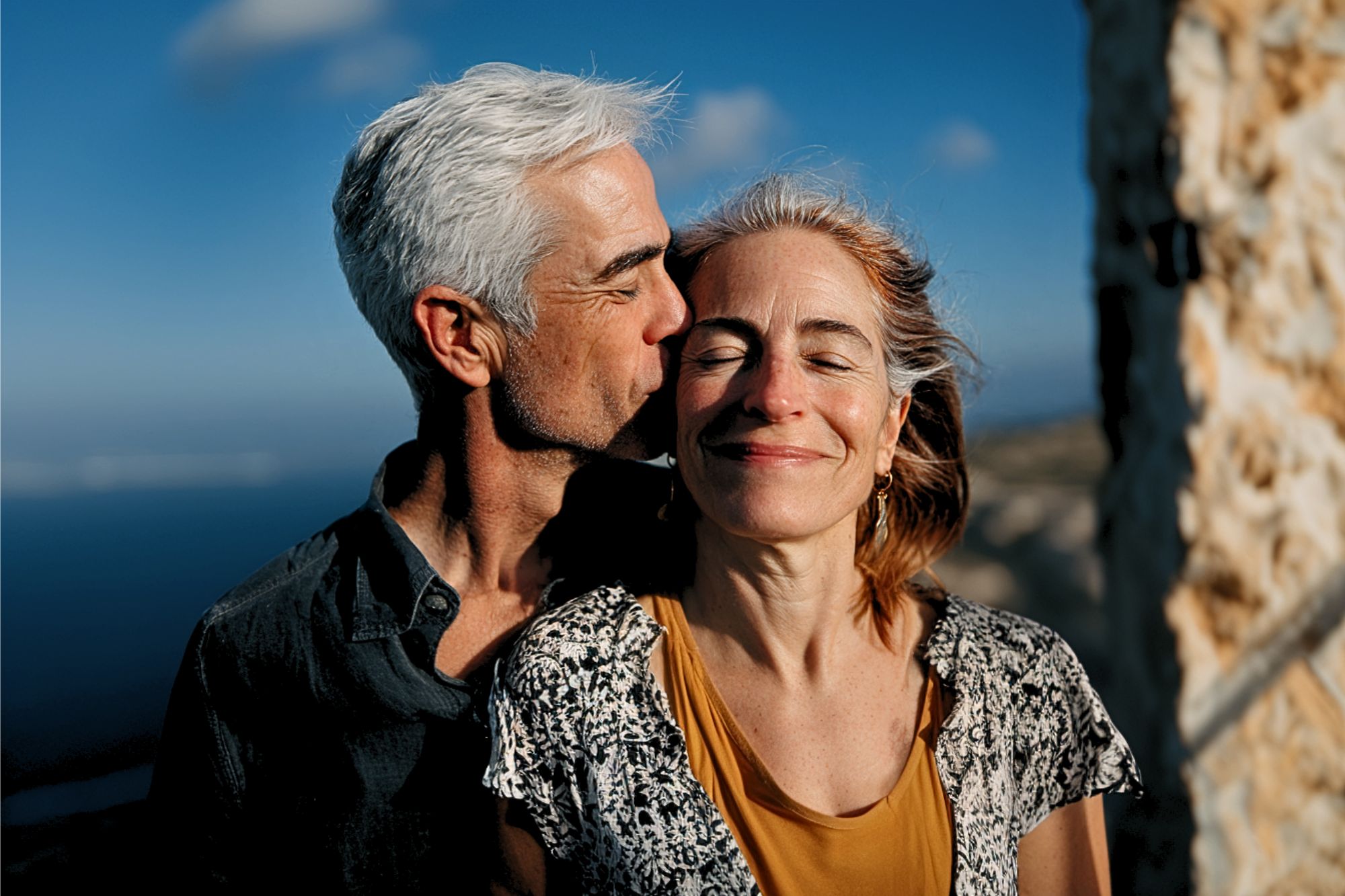 A man with gray hair gently kisses a smiling woman on the forehead. They stand outdoors by a stone wall, bathed in sunlight, with a blue sky and ocean in the background.