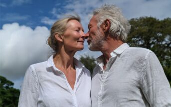 An older couple in white shirts stand close outdoors, touching noses affectionately and smiling, with a blue sky and trees in the background.
