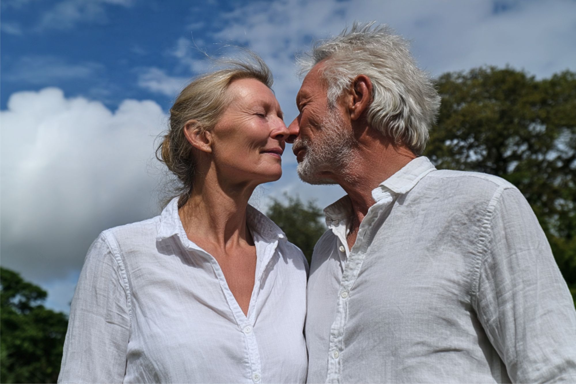 An older couple in white shirts stand close outdoors, touching noses affectionately and smiling, with a blue sky and trees in the background.