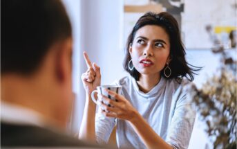 A woman with short dark hair, wearing hoop earrings and a light sweater, is sitting and holding a mug. She is gesturing with her other hand, pointing upwards, and appears to be engaged in conversation with a person blurred in the foreground.