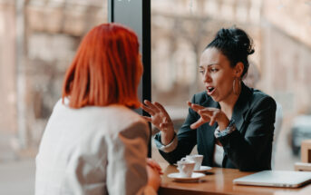 If you lack these 10 communication skills, it’s holding you back in life Two women sit across from each other at a wooden table in a café, engaging in conversation. One woman with red hair listens while the other gestures expressively. They each have a small cup of coffee in front of them.