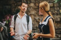 A man and a woman stand outdoors, engaged in conversation. The man gestures with his hands while talking, wearing a white shirt and backpack. The woman, in a blue dress with white accents, holds a cup. They are in a stone-walled setting.