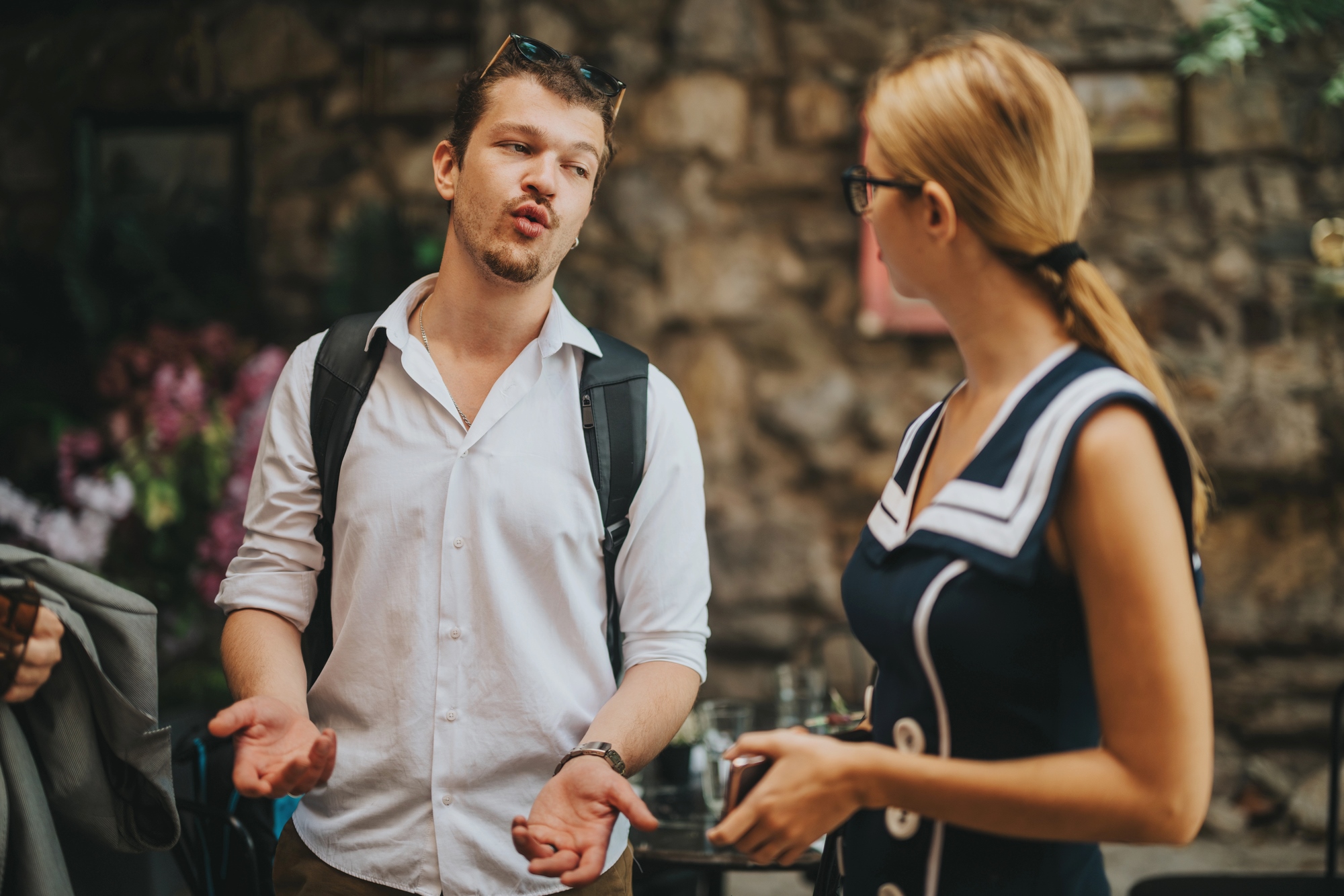 A man and a woman stand outdoors, engaged in conversation. The man gestures with his hands while talking, wearing a white shirt and backpack. The woman, in a blue dress with white accents, holds a cup. They are in a stone-walled setting.