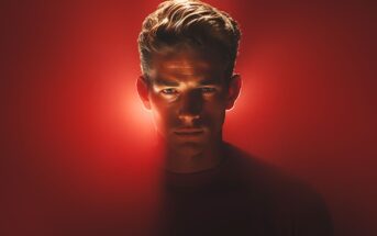 A man with light hair stands against a red background, illuminated dramatically from the front. His face is serious and partially in shadow, with a glow outlining his head.