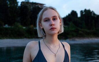 A young woman with short blonde hair gazes at the camera, wearing a dark swimsuit and a necklace. She stands by a calm body of water, with a blurred background of trees and a building at dusk.
