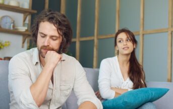 A man with a thoughtful expression sits on a couch, resting his chin on his hand, wearing a light shirt. A woman in a white top sits behind him, leaning back with her arms crossed, holding a blue cushion. The setting is a cozy living room.