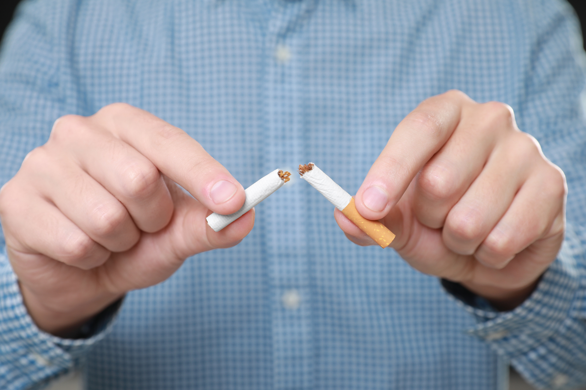 A person in a blue checkered shirt breaks a cigarette in half, symbolizing quitting smoking or resistance to tobacco use. The focus is on their hands and the broken cigarette.