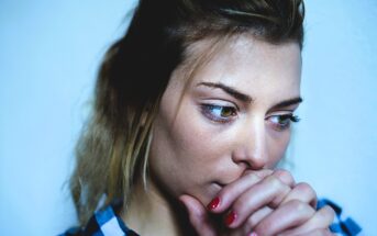 How To Accept Your Anxiety: 14 No Nonsense Tips A woman with light brown hair gazes downward thoughtfully, resting her chin on her clasped hands. She wears a checkered shirt and has red nails, set against a plain blue background.