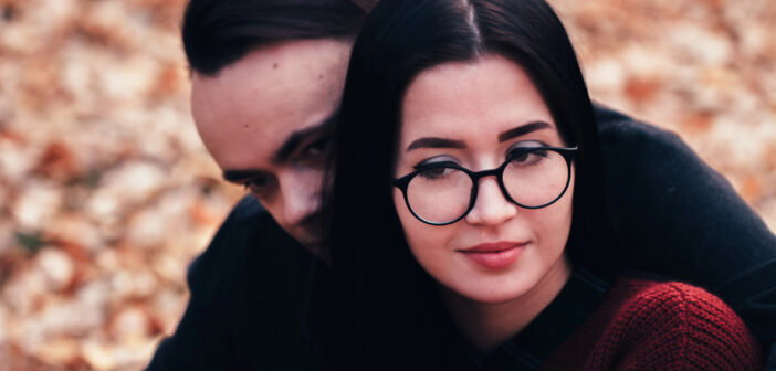 A woman with glasses and dark hair, wearing a red sweater, is sitting in front of a man with dark hair. They are outdoors with autumn leaves on the ground. The man partially hides his face behind her shoulder.