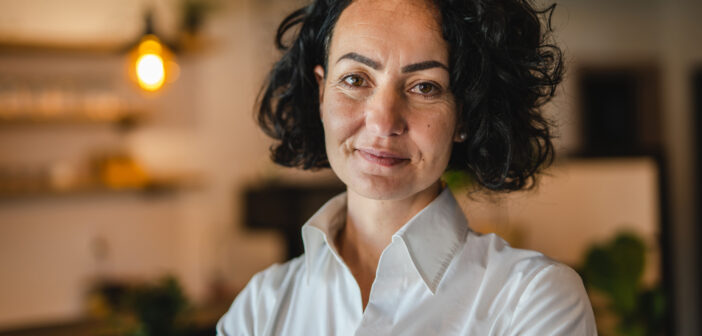 A person with curly hair wearing a white shirt stands indoors with a warm expression. The background is softly lit, featuring a hanging light bulb and blurred shelves.