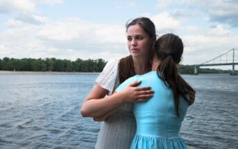 Two women embracing by a river under a partly cloudy sky. One woman faces the camera with a thoughtful expression, wearing a white dress, while the other, in a blue dress, has her back to the camera. A bridge and trees are visible in the background.