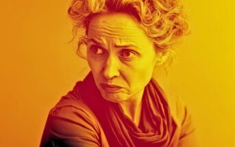 A woman with curly hair and a scarf frowns and looks to the side, her expression serious or concerned. The image is tinted with warm orange and yellow colors.