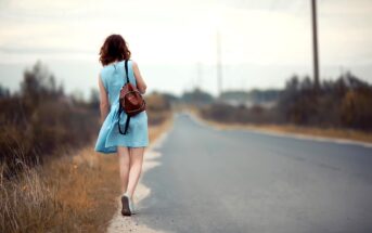 A woman in a light blue dress walks down a rural road, carrying a brown backpack. The scene is serene, with overcast skies and grassy fields on either side of the road.
