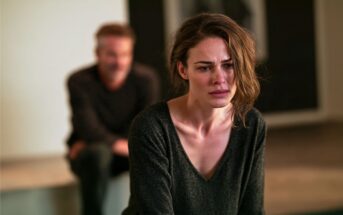 A woman with a distressed expression sits in focus, while a man sits blurred in the background. The scene suggests emotional tension between the two individuals.