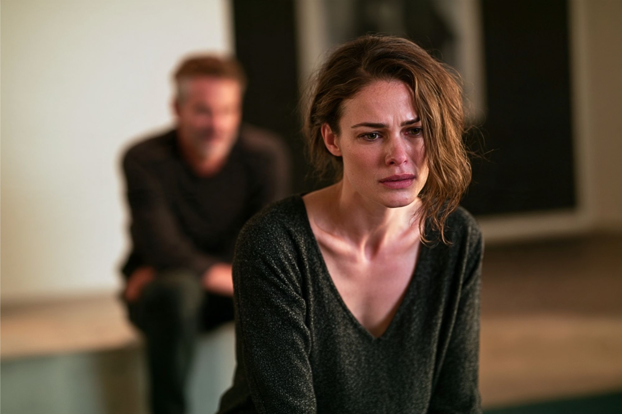 A woman with a distressed expression sits in focus, while a man sits blurred in the background. The scene suggests emotional tension between the two individuals.