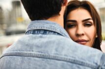 A woman with long brown hair is being embraced by a person wearing a denim jacket, seen from behind. Her face is partially visible over their shoulder, and she has a serene expression. The background is softly blurred.