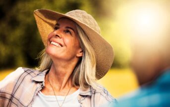 A smiling woman wearing a wide-brimmed straw hat and plaid shirt looks upwards towards sunlight. The background is blurred greenery, and there's a partial view of another person in the foreground. The scene appears joyful and sunlit.