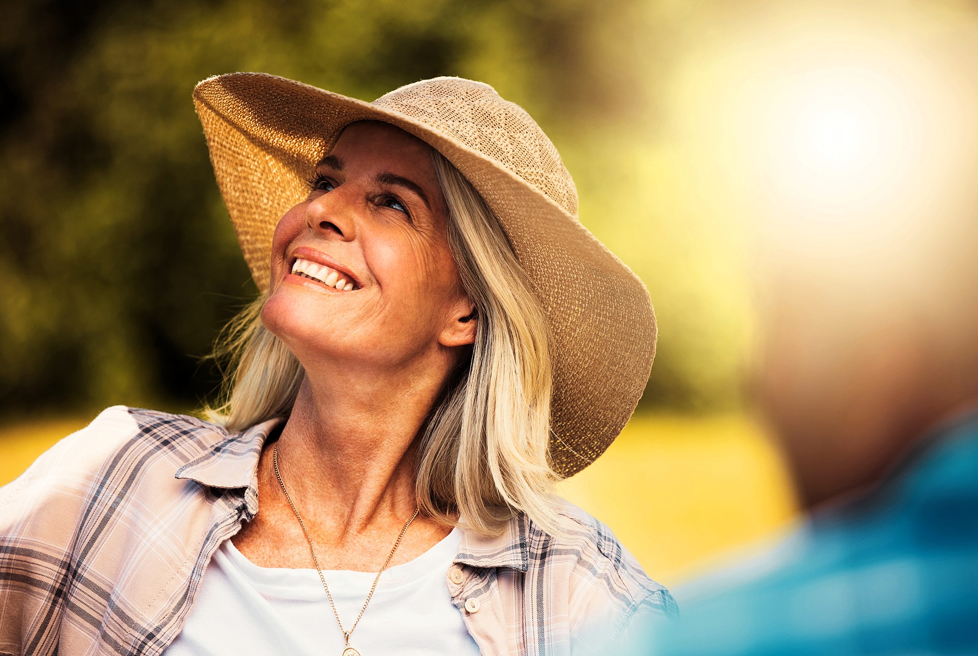 A smiling woman wearing a wide-brimmed straw hat and plaid shirt looks upwards towards sunlight. The background is blurred greenery, and there's a partial view of another person in the foreground. The scene appears joyful and sunlit.