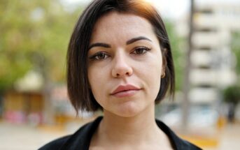 A person with short dark hair and a neutral expression is outdoors with a blurred background featuring trees and a building. They're wearing a black shirt, and soft natural light illuminates their face.