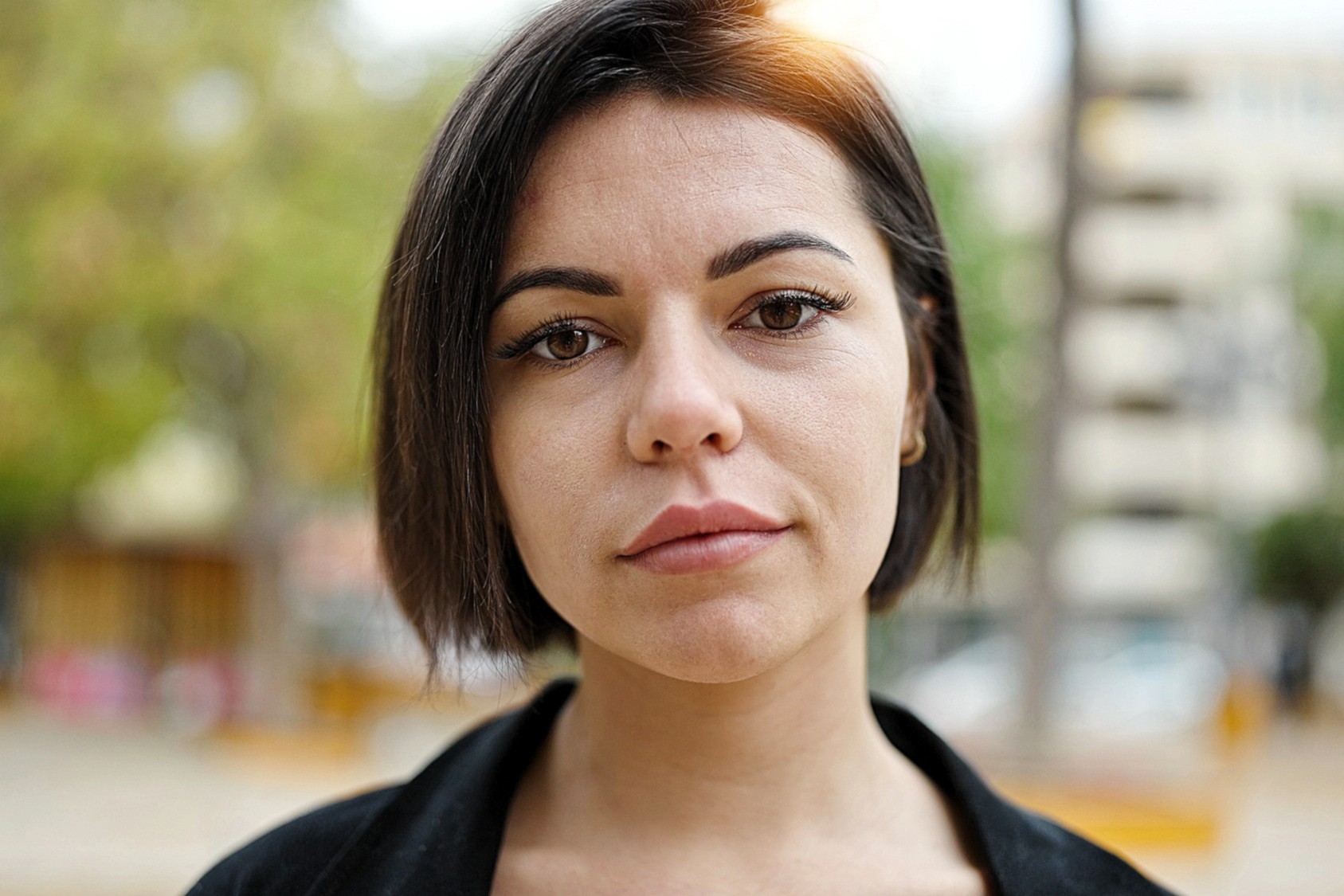 A person with short dark hair and a neutral expression is outdoors with a blurred background featuring trees and a building. They're wearing a black shirt, and soft natural light illuminates their face.