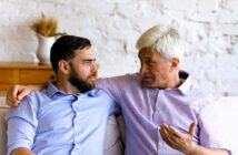 Older man with gray hair and younger bearded man sit closely on a couch, engaging in a conversation. Both wear collared shirts. The older man gestures with his hand while speaking. A vase with wheat is on a wooden cabinet in the background.