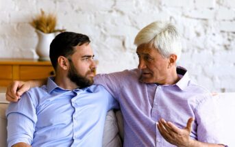 Older man with gray hair and younger bearded man sit closely on a couch, engaging in a conversation. Both wear collared shirts. The older man gestures with his hand while speaking. A vase with wheat is on a wooden cabinet in the background.