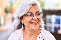 Smiling woman with gray hair and glasses outdoors, wearing a light-colored blouse and cardigan. She looks to the side with a joyful expression. The background is softly blurred, suggesting a street scene.