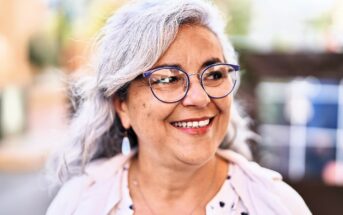 Smiling woman with gray hair and glasses outdoors, wearing a light-colored blouse and cardigan. She looks to the side with a joyful expression. The background is softly blurred, suggesting a street scene.
