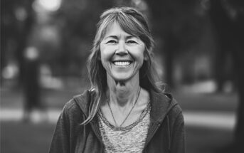 A smiling middle-aged woman with long hair stands outdoors in a park, wearing a casual jacket and t-shirt. The background is blurred, highlighting her cheerful expression. The photo is in black and white.