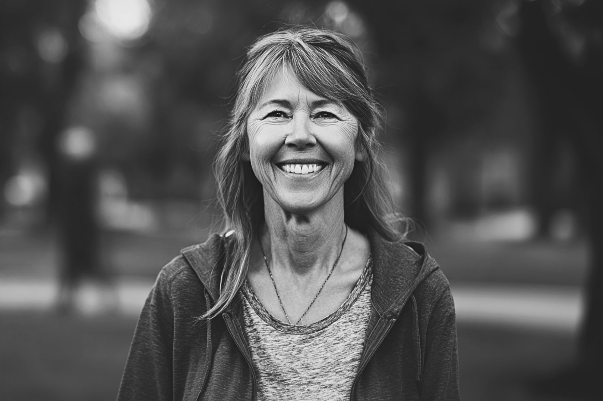 A smiling middle-aged woman with long hair stands outdoors in a park, wearing a casual jacket and t-shirt. The background is blurred, highlighting her cheerful expression. The photo is in black and white.