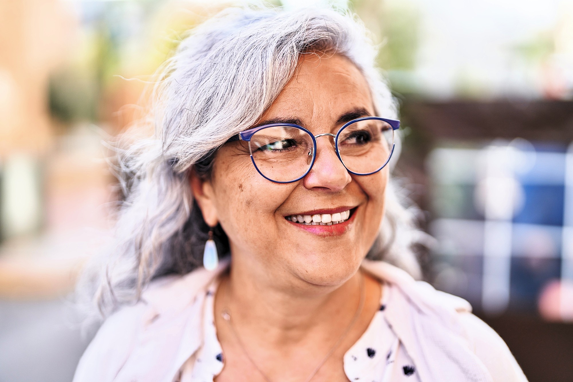 Smiling woman with gray hair and glasses outdoors, wearing a light-colored blouse and cardigan. She looks to the side with a joyful expression. The background is softly blurred, suggesting a street scene.