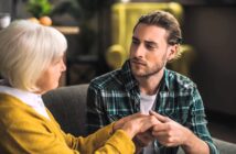 A young man with long hair and a plaid shirt holds the hands of an elderly woman with short white hair and a yellow sweater. They are sitting on a sofa, engaged in a serious conversation, with a blurred background.