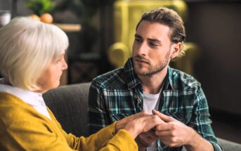 A young man with long hair and a plaid shirt holds the hands of an elderly woman with short white hair and a yellow sweater. They are sitting on a sofa, engaged in a serious conversation, with a blurred background.