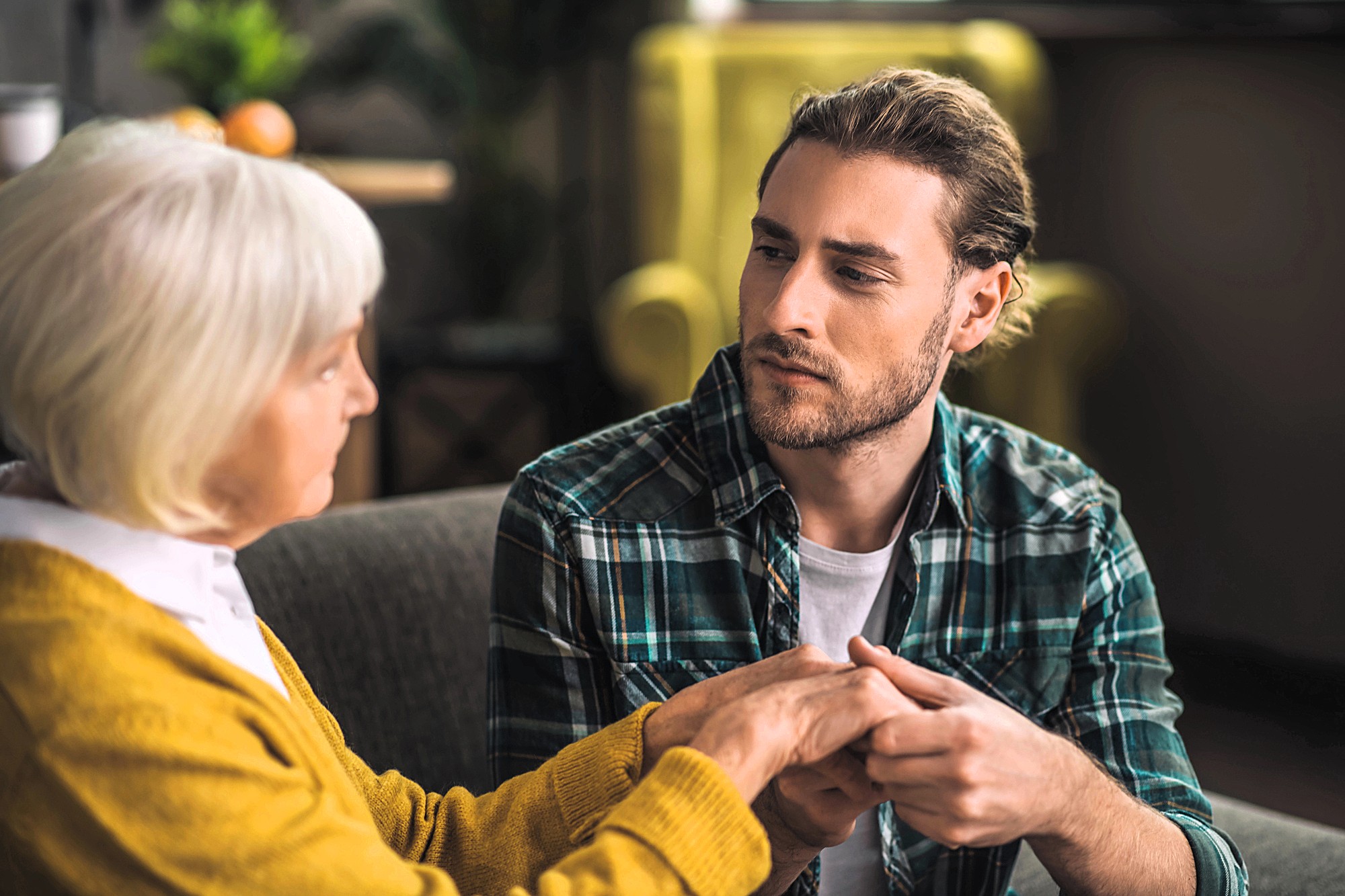 A young man with long hair and a plaid shirt holds the hands of an elderly woman with short white hair and a yellow sweater. They are sitting on a sofa, engaged in a serious conversation, with a blurred background.