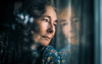 An elderly woman with curly hair gazes pensively out of a window, her face reflected in the glass. She wears a patterned blouse and appears deep in thought or contemplation.
