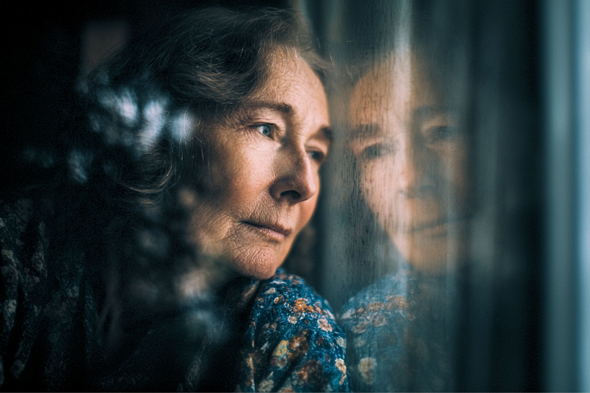 An elderly woman with curly hair gazes pensively out of a window, her face reflected in the glass. She wears a patterned blouse and appears deep in thought or contemplation.