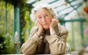 An elderly woman with gray hair sits at a table in a sunlit greenhouse, resting her face on her hands and looking thoughtfully at the camera. She wears a light brown coat and scarf. Green plants surround her.