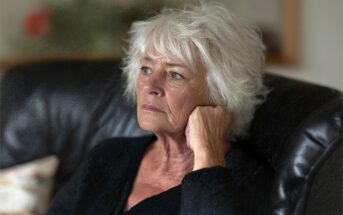 An older woman with white hair sits on a dark couch, resting her head on her hand and looking thoughtfully into the distance. She wears a dark top and appears deep in thought.