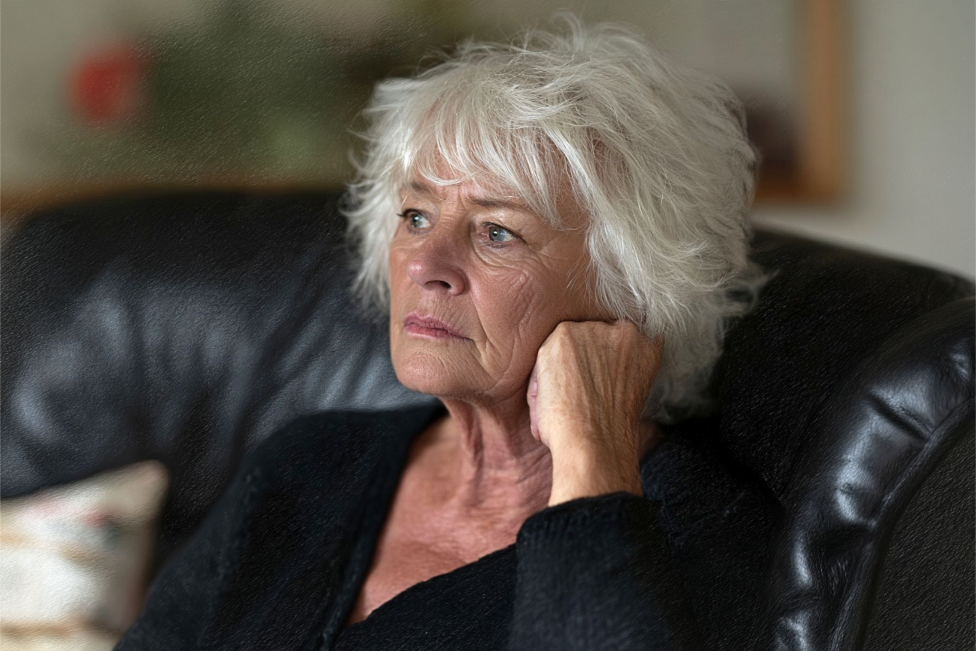 An older woman with white hair sits on a dark couch, resting her head on her hand and looking thoughtfully into the distance. She wears a dark top and appears deep in thought.