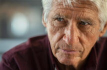 Elderly man with white hair and deep wrinkles wearing a dark maroon shirt, looking pensively to the side. The background is softly blurred, highlighting the introspective expression on his face.