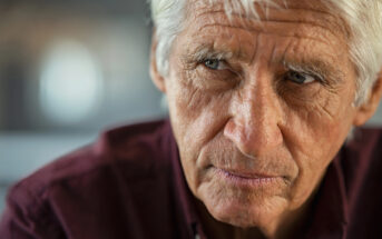 Elderly man with white hair and deep wrinkles wearing a dark maroon shirt, looking pensively to the side. The background is softly blurred, highlighting the introspective expression on his face.
