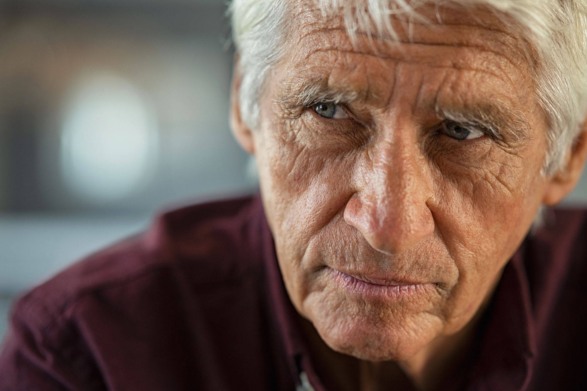 Elderly man with white hair and deep wrinkles wearing a dark maroon shirt, looking pensively to the side. The background is softly blurred, highlighting the introspective expression on his face.