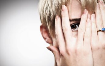 A close-up of a person with short blonde hair gazing through their fingers. Their hands are partially covering their face, with a blue-stoned ring visible on the right hand. The background is a soft gray gradient.
