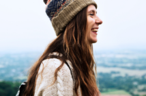 A woman with long hair is smiling, wearing a knit beanie and sweater, standing outdoors. The background is a blurred view of a landscape with greenery and sky.