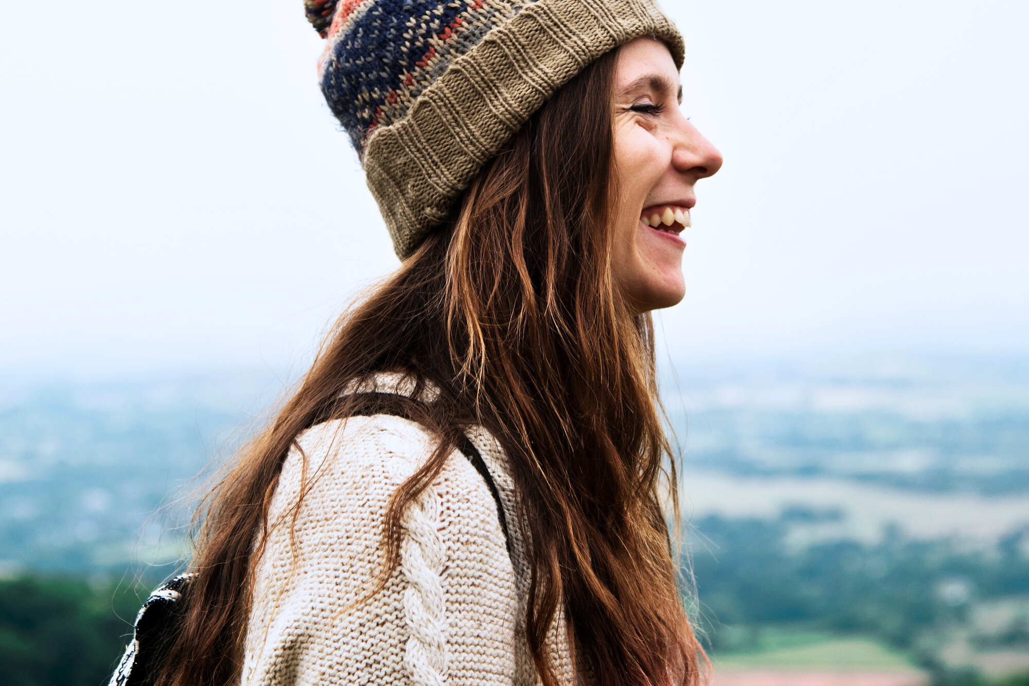 A woman with long hair is smiling, wearing a knit beanie and sweater, standing outdoors. The background is a blurred view of a landscape with greenery and sky.