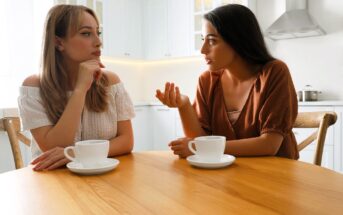 Two women sit at a wooden table in a bright kitchen, engaged in conversation. One gestures with her hand while speaking, and both have cups of coffee in front of them. The kitchen features white cabinets and a stovetop in the background.