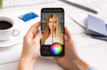 A person holds a smartphone displaying a hair color app. The screen shows a woman's hair before and after color adjustment, with a color wheel and sliders to modify saturation and lightness. A coffee cup, card, and papers are on the table.