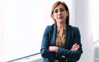 A woman with shoulder-length hair stands confidently in a well-lit room, wearing a dark blazer over a brown top. Her arms are crossed, and she is looking directly at the camera. A large window is visible in the background.